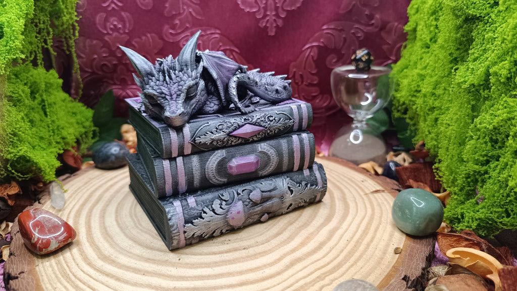 Cajita joyero Eclipse: un dragón dormido enroscado sobre una pila de tres libros antiguos, impresa en 3D y pintada a mano en gris plata y violeta.