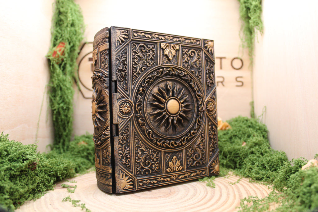 Caja secreta Alborada con forma de libro antiguo, con un sol de rayos abiertos en el centro de la tapa y filigrana alrededor, pintada a mano en dorado sobre fondo oscuro.