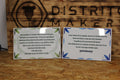 Dos azulejos personalizados de 20 × 30 cm con dedicatorias a los vecinos de la esquina, uno con cenefa de hojas verdes y otro con cenefa de hojas azules, delante del logo de Distrito Makers grabado en madera